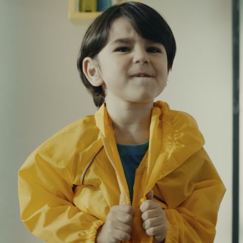 Um menino ajeita sua capa de chuva amarela com suas mãos enquanto morde seus lábios.
