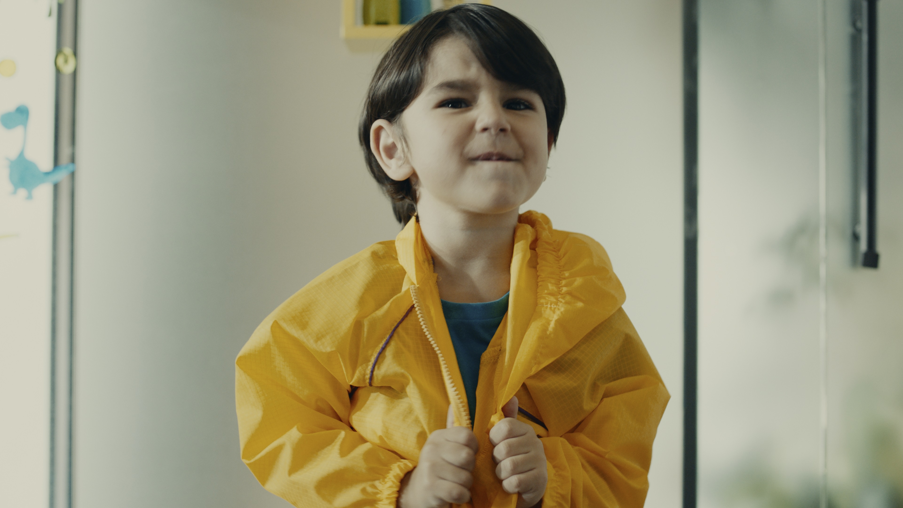 Um menino ajeita sua capa de chuva amarela com suas mãos enquanto morde seus lábios.