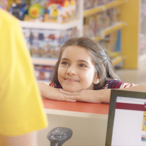 Uma menina apoia a cabeça sobre suas mãos sobre o balcão da loja de brinquedos Ri Happy. Ela olha para o atendente do caixa enquanto sorri.