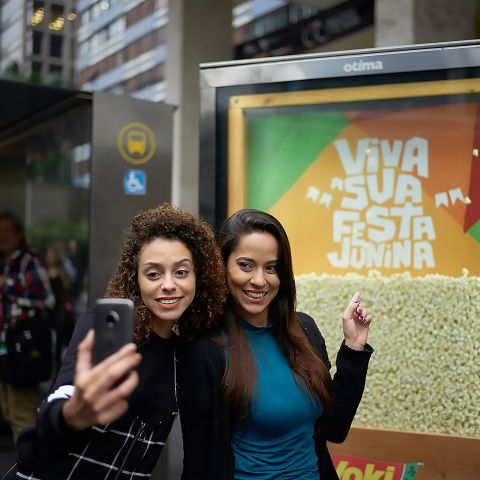 Na imagem, duas mulheres posam para uma selfie em frente ao display publicitário Yoki em um ponto de ônibus. Dentro do display há pipoca pronta com a frase: Viva sua festa junina.