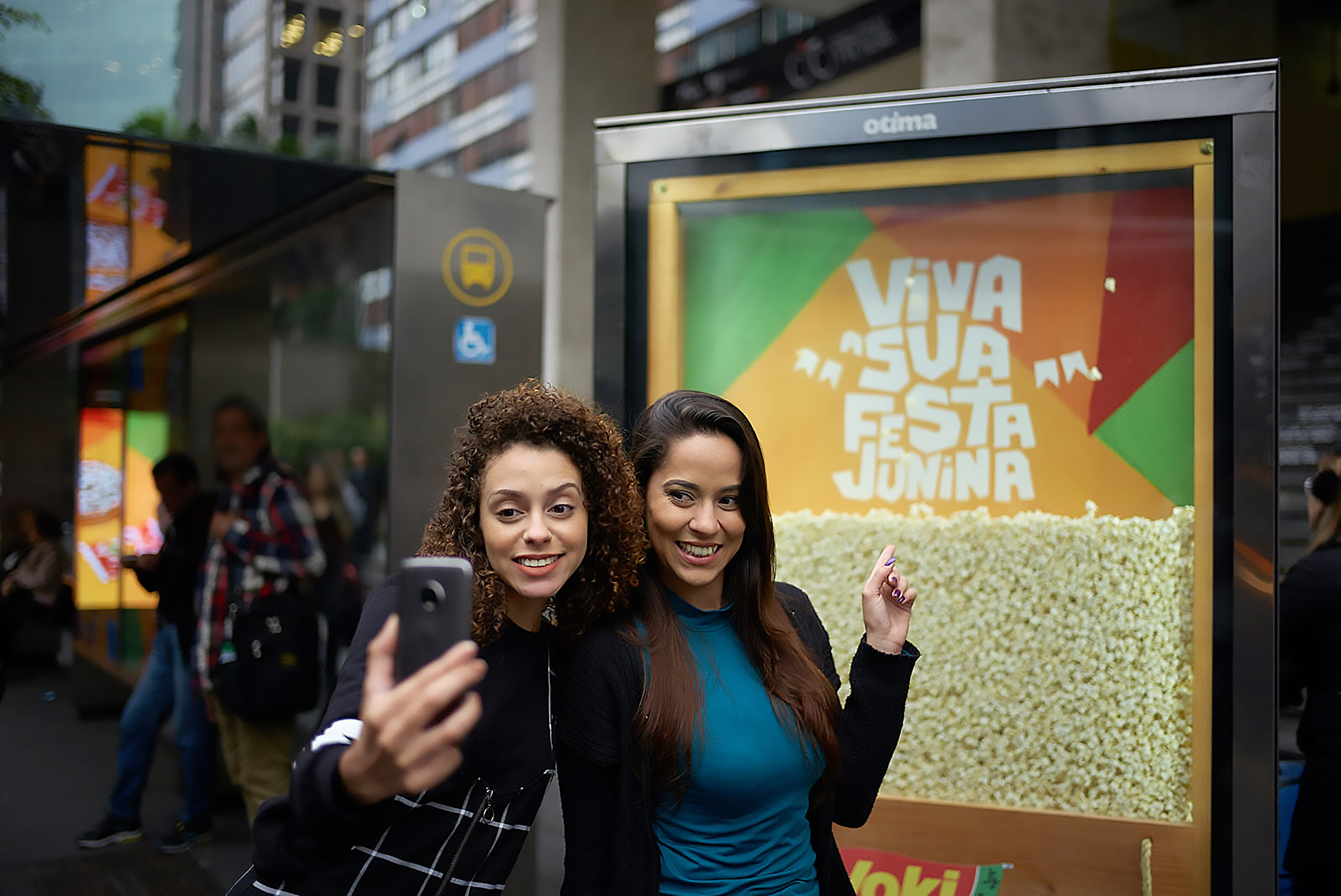 Na imagem, duas mulheres posam para uma selfie em frente ao display publicitário Yoki em um ponto de ônibus. Dentro do display há pipoca pronta com a frase: Viva sua festa junina.