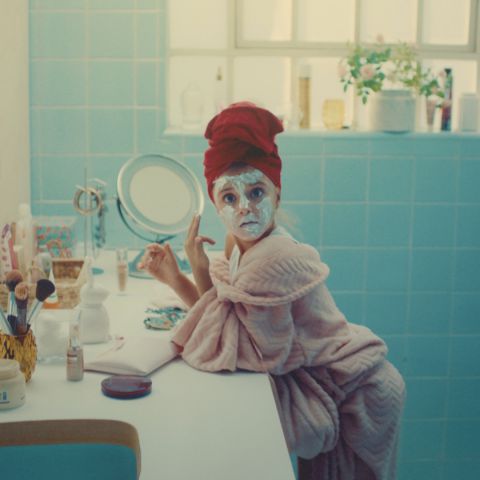 Uma menina faz cara de surpresa quando é pega vestindo roupão de banho e toalha na cabeça enquanto passa uma máscara de beleza no rosto