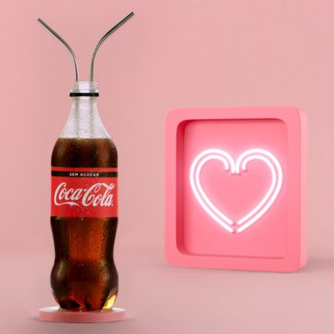 Na imagem, há uma garrafa de Coca-Cola com dois canudos dentro. Ao lado, um coração rosa com neon branco.