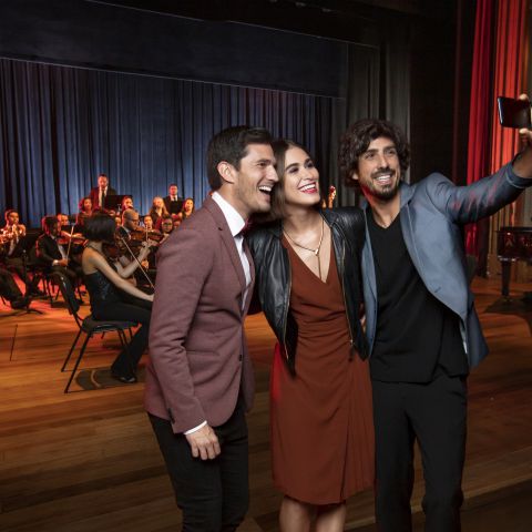 Uma mulher e dois homem tiram uma selfie em um concerto musical. O homem à direita tem cabelos pretos, barba e veste um blêizer cinza, camiseta preta e calça preta. A mulher no centro usa vestido marrom, com jaqueta de couro preta e um colar dourado. O homem à esquerda veste um blêizer de cor vermelho-escuro, camisa branca e calça preta. No fundo da imagem há instrumentistas tocando violinos à esquerda e um pianista à direita.