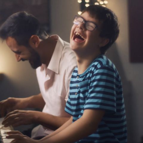 Um menino sorri enquanto toca piano ao lado do seu pai