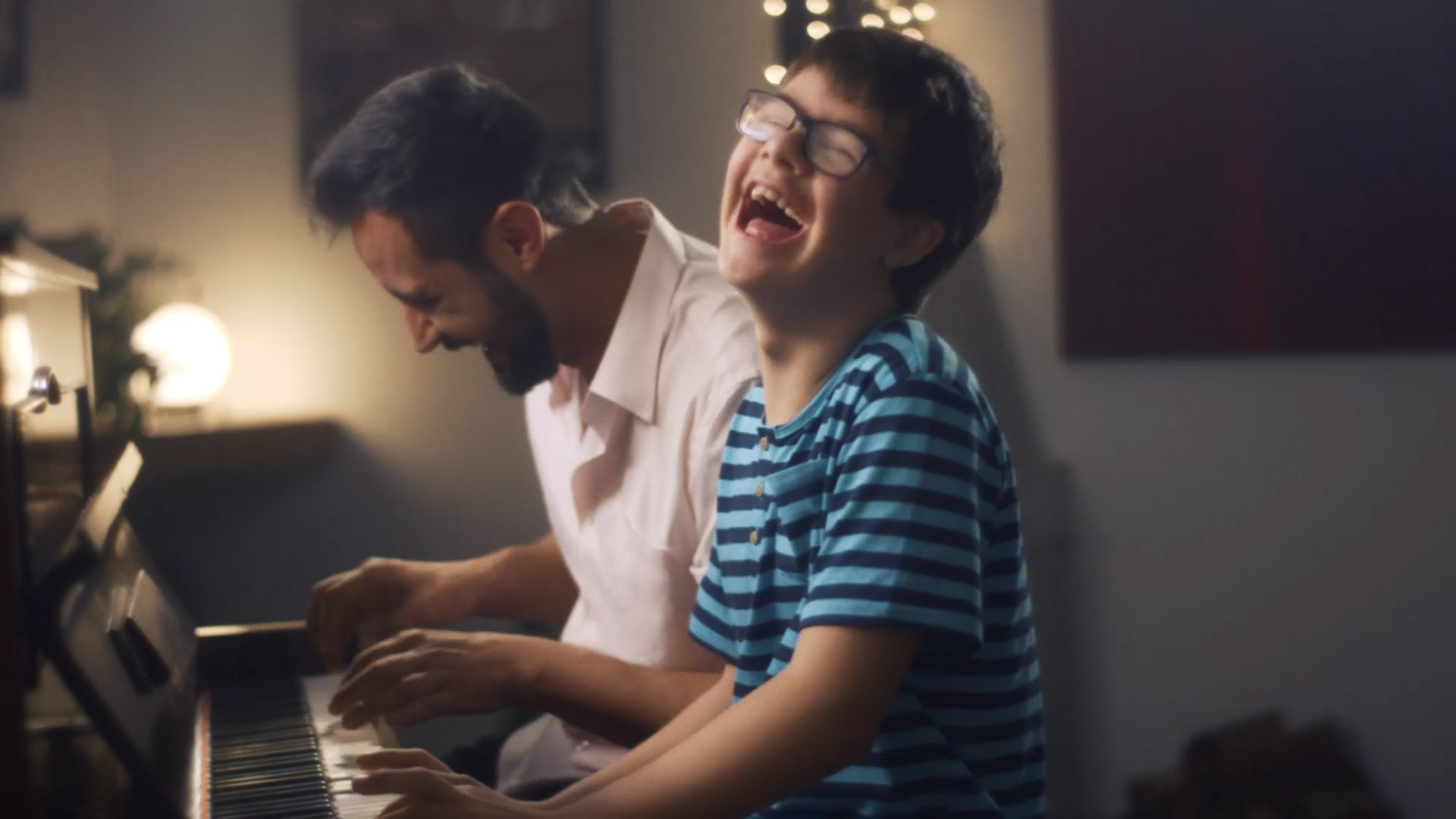 Um menino sorri enquanto toca piano ao lado do seu pai