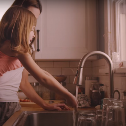 Mãe e filha juntas em frente a bancada da cozinha, usando a torneira ligada.