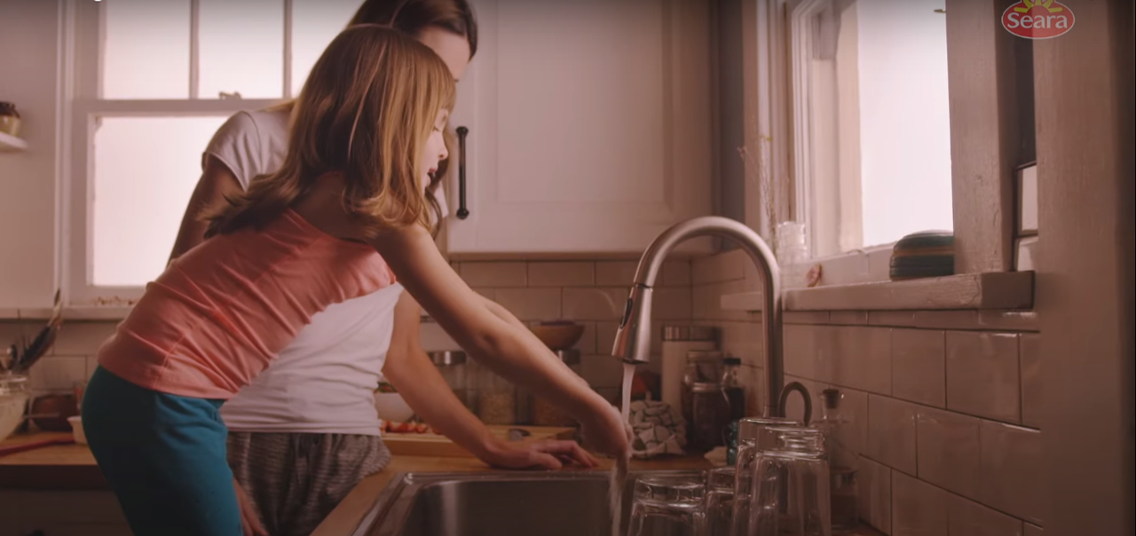 Mãe e filha juntas em frente a bancada da cozinha, usando a torneira ligada.