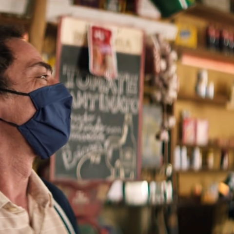 Na imagem, a cena de um comerciante, atrás do balcão de sua loja, usando máscara de proteção e com uma expressão de sorriso.