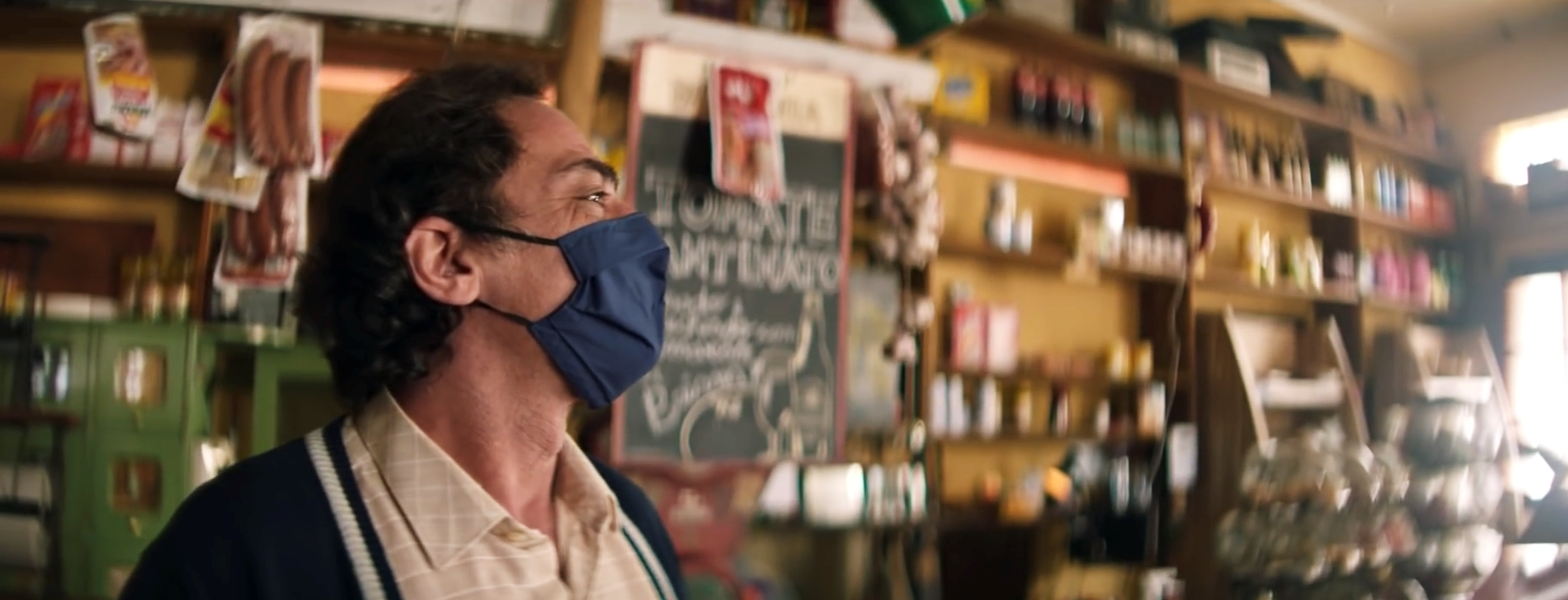 Na imagem, a cena de um comerciante, atrás do balcão de sua loja, usando máscara de proteção e com uma expressão de sorriso.