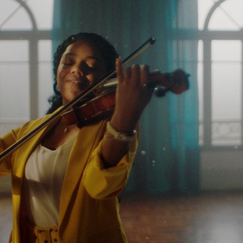 Na imagem, uma mulher vestida de blusa amarela toca violino com leve sorriso no rosto.