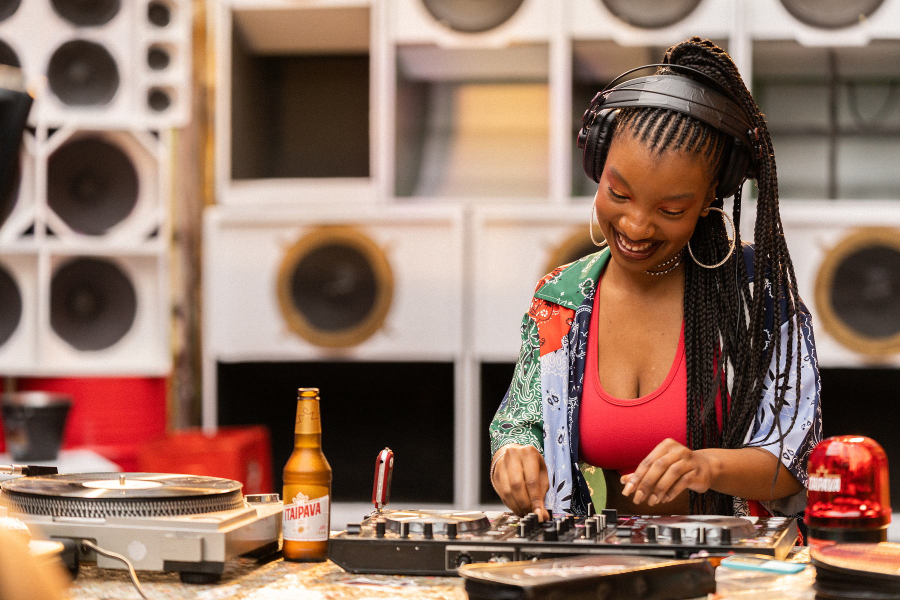 Na imagem encontramos uma mulher negra tocando em um painel de DJ ao fundo varias caixas de som grande. Pequena, em preto e branco