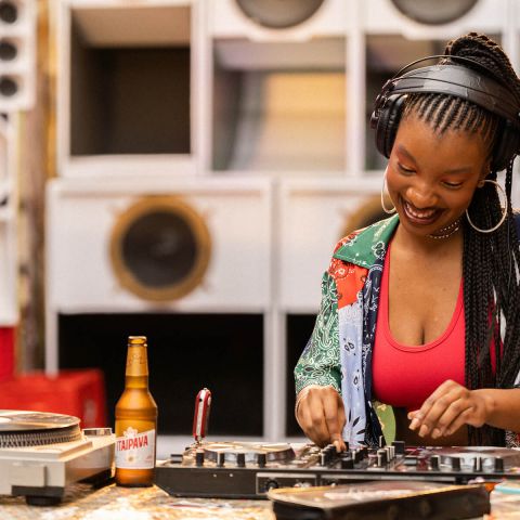 Na imagem encontramos uma mulher negra tocando em um painel de DJ ao fundo varias caixas de som grande